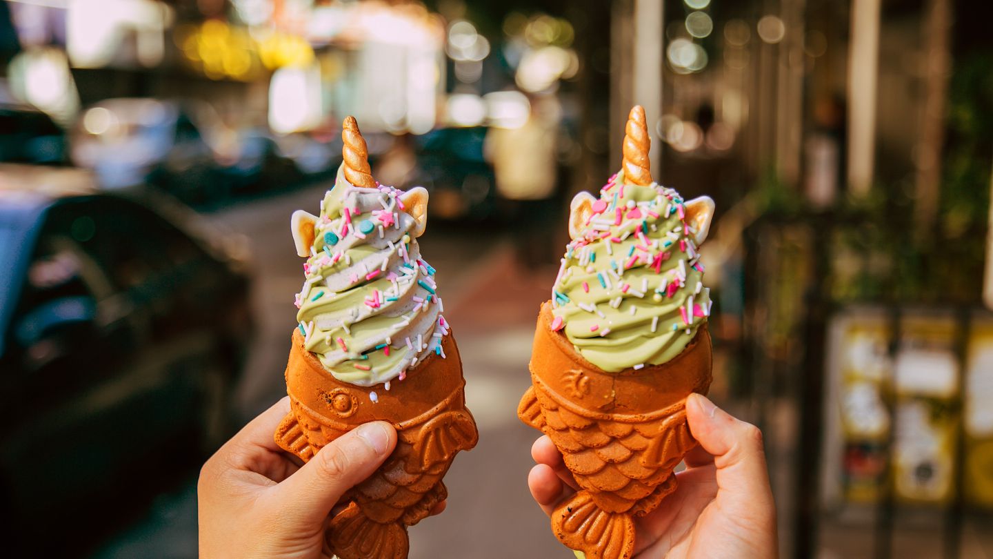 A person holds two ice cream cones, one in each hand. The cones are shaped like fish with their mouths wide open and ice cream has been piped into the mouth. The ice cream in both cones looks like soft serve and is green. They are topped with colorful sprinkles and what appear to be unicorn horns and ears.