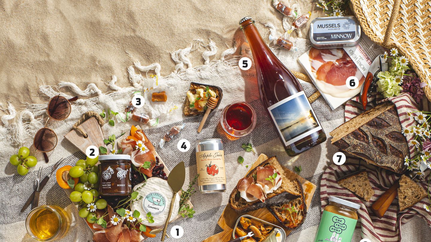 Picnic ingredients laid out on a stripped blanked set on some sand. From left, there are grapes, fruit preserves, camambrie cheese, apple juice in a can, a bottle of wine, a cutting board with some bread topped with cured meat, a tin of mussels, a jar of peanut butter, a loaf of bread, and a package of cured meat and another tin of mussels.