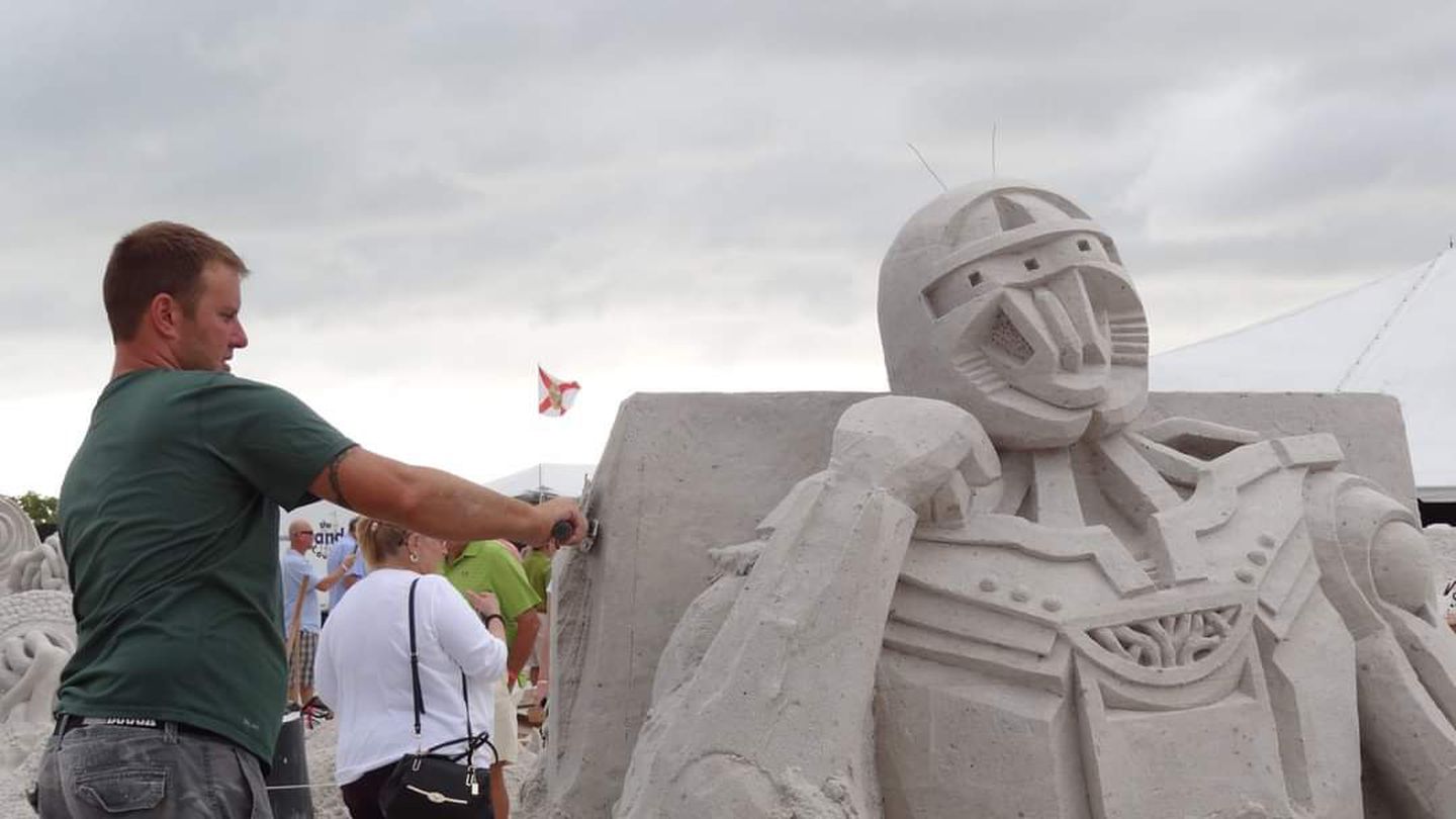 A man uses a trowel to work on the left side of a sand sculpture. It looks like it's a knight or maybe a Transformer.